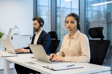 Two customer service representatives work in an office setting, each wearing a headset and using a laptop.