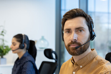 A professional man wearing a headset, possibly a call center representative, looks intently at the camera in an office setting. Another employee is visible in the background.