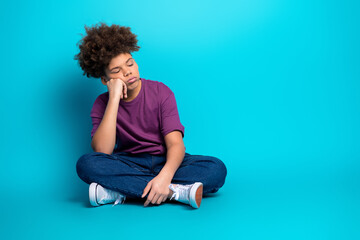 Young boy sitting cross legged on a blue background, feeling sleepy with head resting on hand
