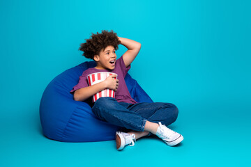 Joyful boy sitting comfortably on a bean bag chair eating popcorn against a bright blue background