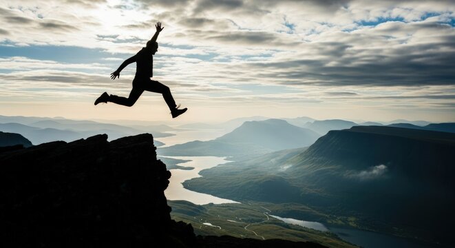 Silhouette of a person jumping off a cliff with mountains and a cloudy sky in the background scenery view