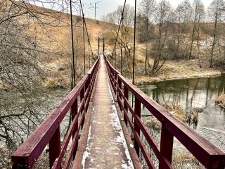 A narrow, wooden suspension bridge spans across a small river, supported by cables. The bridge has red rails and a snow-dusted walkway. Surrounding the bridge, leafless trees and patches of dry grass 