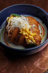 Gourmet croissant with creamy corn filling in blue bowl – elegant food presentation on rustic copper background. vertical close up