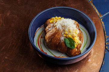 Gourmet croissant with creamy corn filling and parmesan cheese in blue bowl – elegant food presentation on rustic copper background