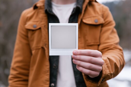 Person Holding Blank Polaroid Frame Outdoors, Wearing Casual Jacket in Nature