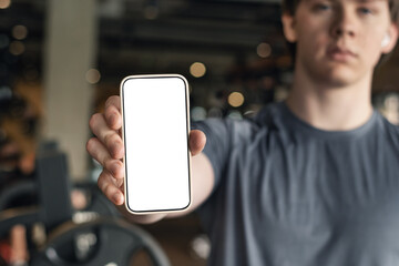 Athlete standing and displaying smartphone with white blank screen in gym setting. Mock up. Space for design.