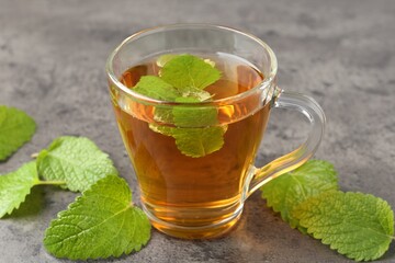 Aromatic lemon balm tea in glass cup and fresh leaves on gray textured table, closeup