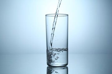 Pouring water into glass at mirror table against light grey gradient background, closeup