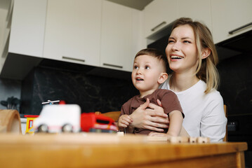 Smiling mother and her cheerful son sitting together at the kitchen table, enjoying playful time surrounded by toy cars and trains.