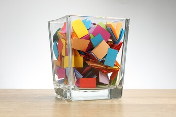 Jar with colorful pieces of paper on wooden table against light background