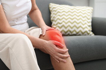 Senior woman suffering from pain in knee on sofa at home, closeup
