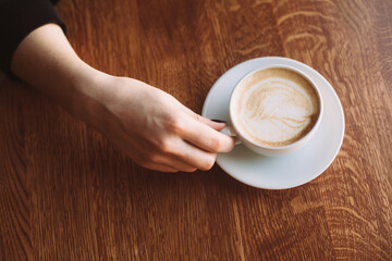 A hand delicately holds a ceramic cup filled with latte on a wooden table.