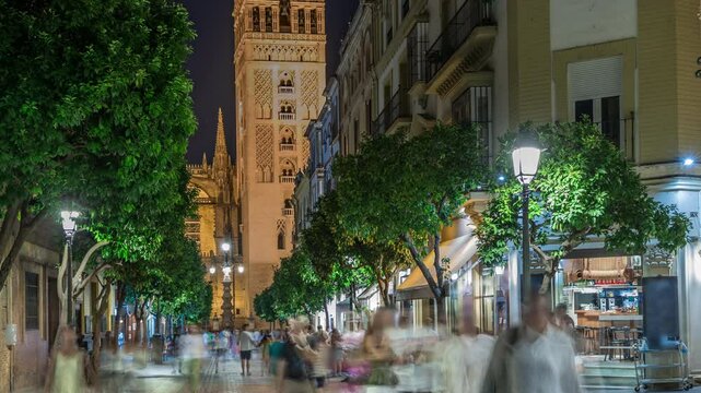 Night timelapse of Barrio Santa Cruz in Seville, Spain, with Giralda tower illuminated in the background. Tourists and locals walk along the charming cobblestone streets lined with cafes and shops.