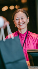 A smiling woman in a bright pink blouse stands behind a counter, engaging with a customer. She holds a black shopping bag, showcasing a welcoming atmosphere.
