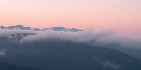 Fototapeta premium Aerial view of mountain range shrouded in fog under a soft pink and purple sky at dawn or dusk