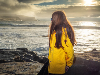 Girl tourist in yellow jacket enjoys stunning nature scenery with rough stone coast, powerful ocean...