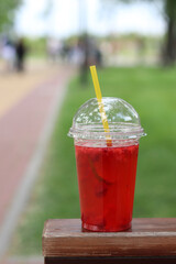 Lemonade in a disposable plastic cup, takeaway drink. Refreshing drink against the background of a park in summer. Pink lemonade with raspberries and lime. Close-up of a drink in a disposable cup