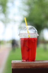 Lemonade in a disposable plastic cup, takeaway drink. Refreshing drink against the background of a park in summer. Pink lemonade with raspberries and lime. Close-up of a drink in a disposable cup