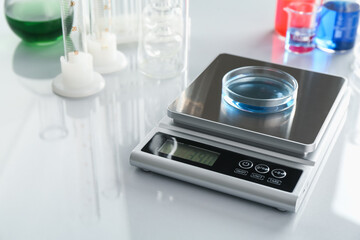 Scale with Petri dish of sample and other laboratory glassware on white mirror table, closeup