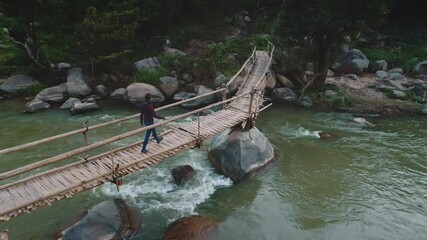 Person walks across a rustic wooden bridge spanning a flowing river, surrounded by lush greenery and large rocks. The scene captures a tranquil moment in a natural setting