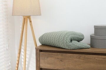 Green woolen blanket, boxes on wooden chest of drawers and lamp near white wall indoors, closeup