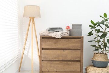 Woolen blankets on wooden chest of drawers, lamp and houseplant near white wall indoors