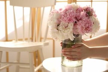 Woman putting beautiful peonies into vase at white table indoors, closeup. Space for text