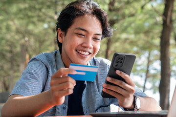 Happy Asian Young businessman hold blue creditcard and phone purchasing online shopping in outdoor cafe.