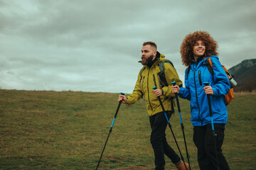 Hikers walking on green meadow with trekking poles and backpacks in cloudy day