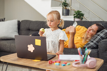 Young blond European girl eating apple with laptop on table. Mother woman relaxing on couch using smartphone. Home environment.