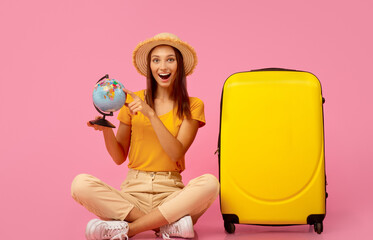 Emotional girl with globe sitting next to yellow luggage over pink background