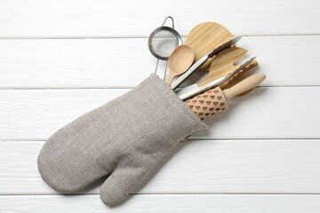 Different kitchen utensils in oven mitten on white wooden table, top view
