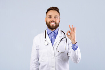 Fototapeta premium Portrait of happy young doctor in uniform showing okay gesture on grey studio background. Professional physician recommending medical treatment or clinic. Healthcare concept
