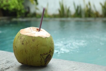 Green coconut with refreshing drink and straw near swimming pool outdoors, closeup. Space for text
