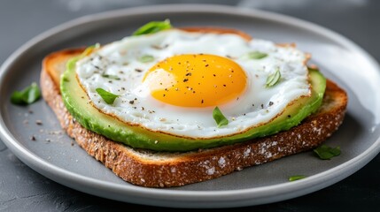 A delicious breakfast plate featuring a perfectly cooked sunny side up egg resting on creamy avocado, served on toasted whole grain bread with a sprinkle of seasoning.
