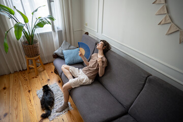 Exhausted tired woman fanning herself on sofa suffering from heat with cat lying nearby struggling in summer temperatures without AC. Animal distress, dehydration exhaustion, seasonal hardship 