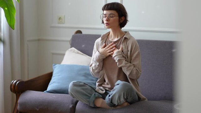Young woman meditating with deep breaths hands on chest sit in lotus position to calm mind at home. Mindfulness stress relief, spiritual controlled breathing, inner emotional regulation, zen routine