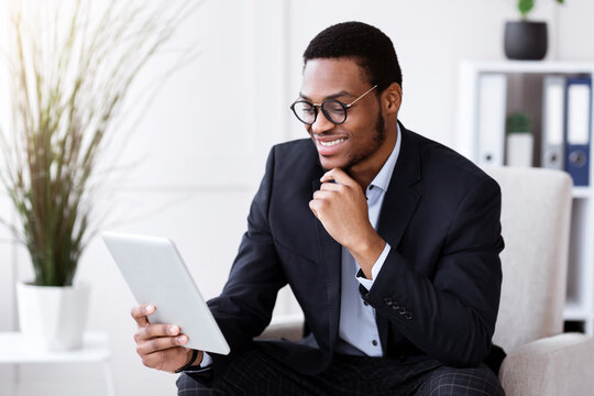 Cheerful black businessman holding digital tablet, office interior, empty space. Smiling african american young entrepreneur using digital pad while working in his office, surfing on internet