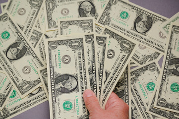 A hand holds several one dollar bills while others are scattered on a flat surface.