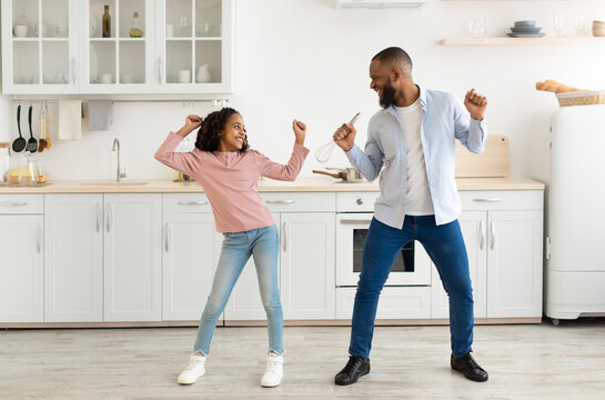 Playful Mood. Excited African American father and joyful girl dancing while cooking together at kitchen, fooling around, cheerful man holding whisk, spending free time with cute daughter