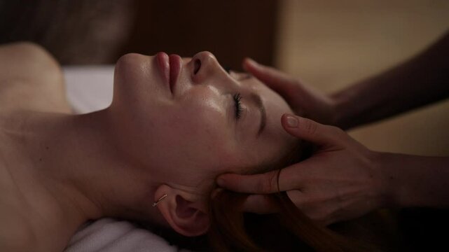 Professional masseuse massaging temples of young relaxed woman, lying with closed eyes on massage table in spa salon. Close-up hands of masseur performing head massage for resting female, slow motion.