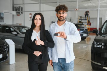 Happy customer showing new car key with saleswoman gesturing thumbs up in dealership