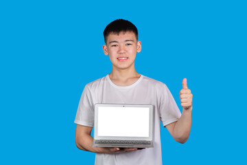 Smiling asian young man teenager in white T-shirt holding modern laptop with blank white screen and showing thumb-up gesture against blue background