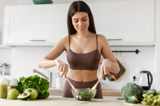 Healthy food. Fit lady making fresh salad, mixing vegetables in bowl in kitchen interior. Dieting and weight loss recipes concept