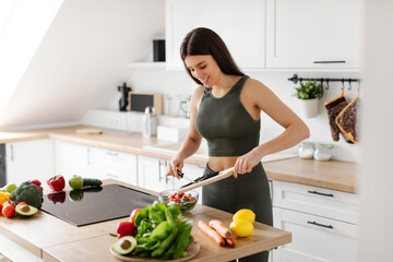 Nutrition tips. Young lady in sports clothes preparing vegetable salad at home in modern kitchen, adding cucumber slices to bowl, free space