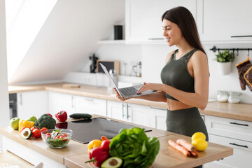 Fit lady using laptop and searching healthy nutrition advices or recipes for slimming, planning menu online standing in kitchen. Weight loss concept
