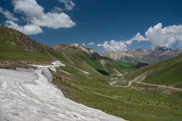 Wide angle landscape of a winding road going through a highland mountain pass surrounded by high peaks with glaciers. Summer day, blue sky with white clouds. Kyrgyzstan, Naryn Region, Central Asia.