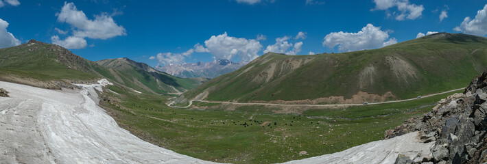 Wide angle landscape panorama of a winding road going through a highland mountain pass, high peaks with glaciers in the background. Summer day, blue sky with white clouds. Kyrgyzstan, Naryn Region.