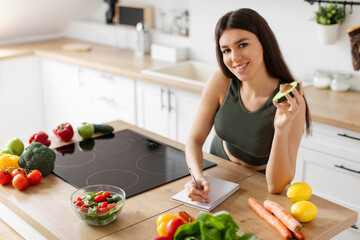 Slim European lady in sportswear writing down healthy recipes or daily ration diet, standing in...