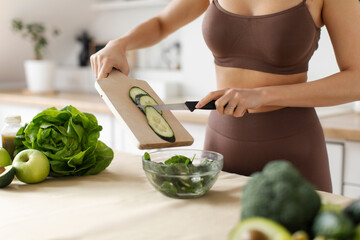 Woman in sportswear adding slices of cucumber to bowl with salad, cooking on table surrounded by...
